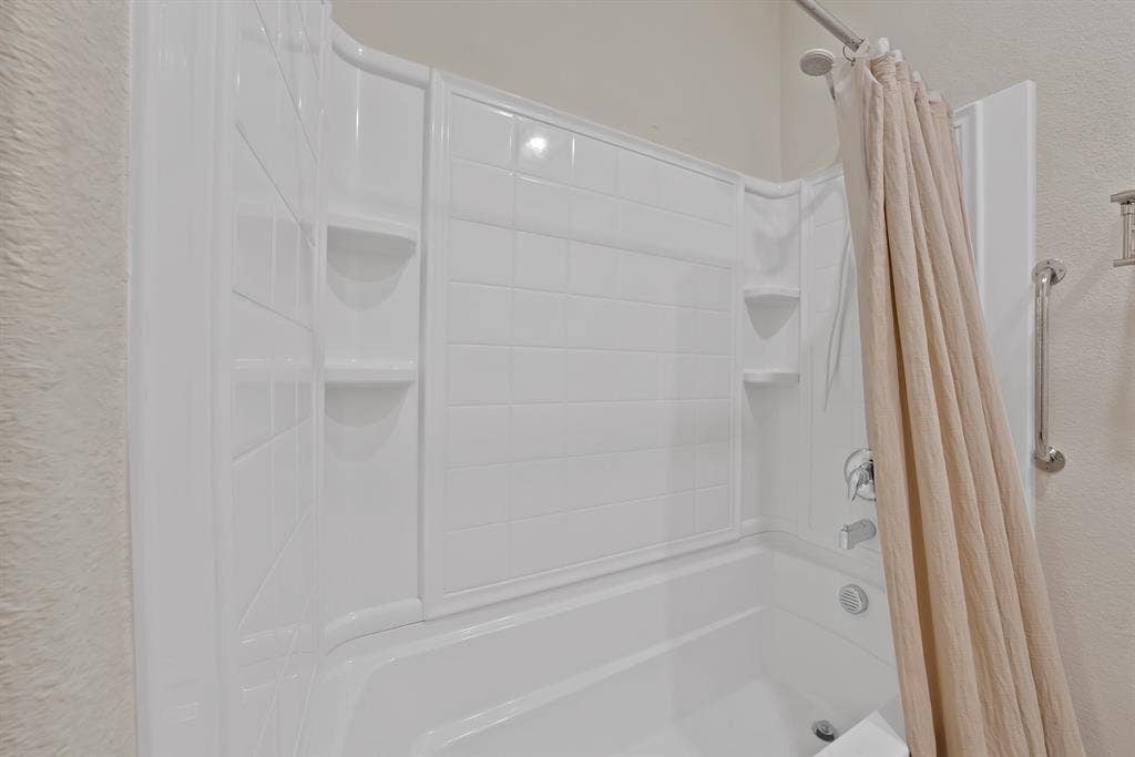228 Foxhunter Street, Fort Worth, TX 76131 Bathtub with white wall surround featuring integrated shelving, a shower head, and a polished chrome grab bar
