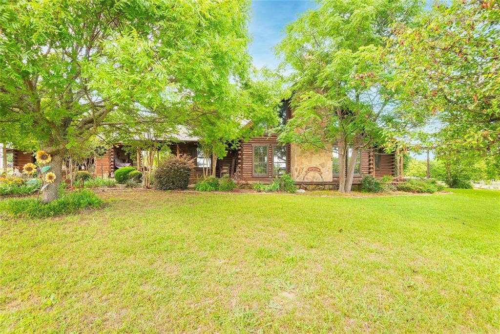 8685 FM 1827, Anna, TX 75409 Log cabin exterior featuring horizontal timber siding, a prominent natural stone chimney, covered front porch, and mature shade trees set upon a manicured lawn