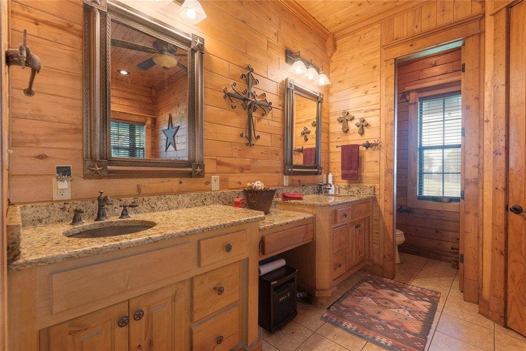8685 FM 1827, Anna, TX 75409 Dual-sink vanity with natural stone countertops, under-mount sinks, wood cabinetry, and framed mirrors with integrated lighting, finished with tile flooring and wood-paneled walls