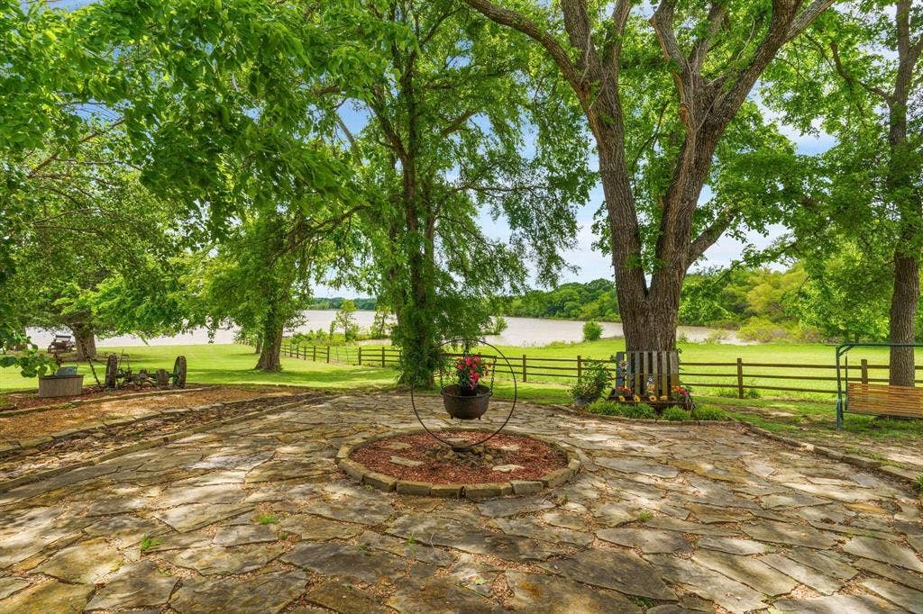 8685 FM 1827, Anna, TX 75409 Flagstone patio featuring a circular stone inlay, mature hardwood trees, grass lawn, wood-post fencing, and water frontage