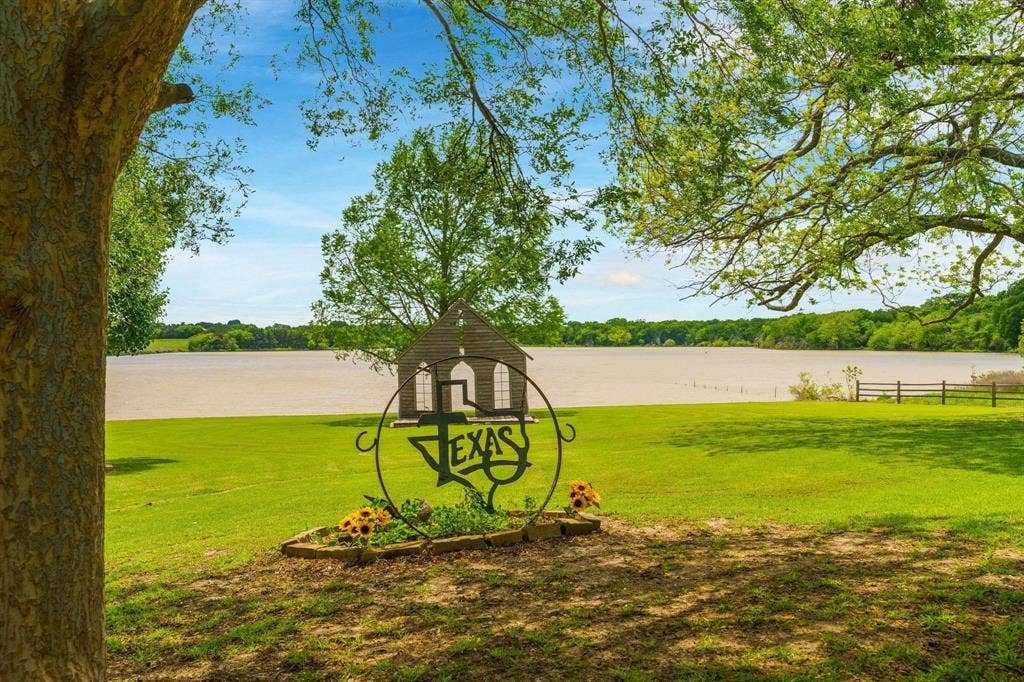 8685 FM 1827, Anna, TX 75409 Expansive waterfront acreage featuring a manicured lawn, mature trees, a metal-framed garden bed, and a wooden open-air structure