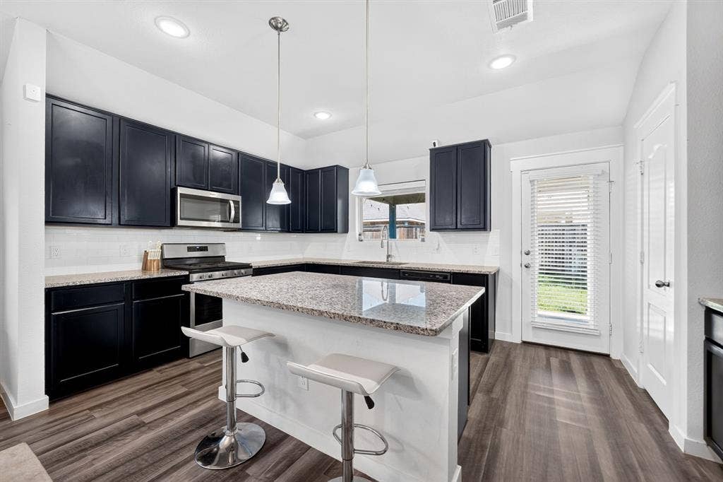 5534 Yarborough Drive, Forney, TX 75126 Modern kitchen featuring black cabinetry with stainless steel appliances, granite countertops, a white kitchen island with seating, pendant lighting, recessed ceiling lights, and wood-finish flooring throughout