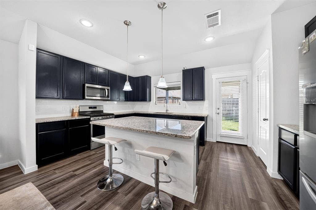 5534 Yarborough Drive, Forney, TX 75126 Modern kitchen featuring black cabinetry with stainless steel appliances, white quartz countertops, a large waterfall-edge island with seating for two, pendant lighting, recessed ceiling lights, and wide-plank wood-finish flooring throughout