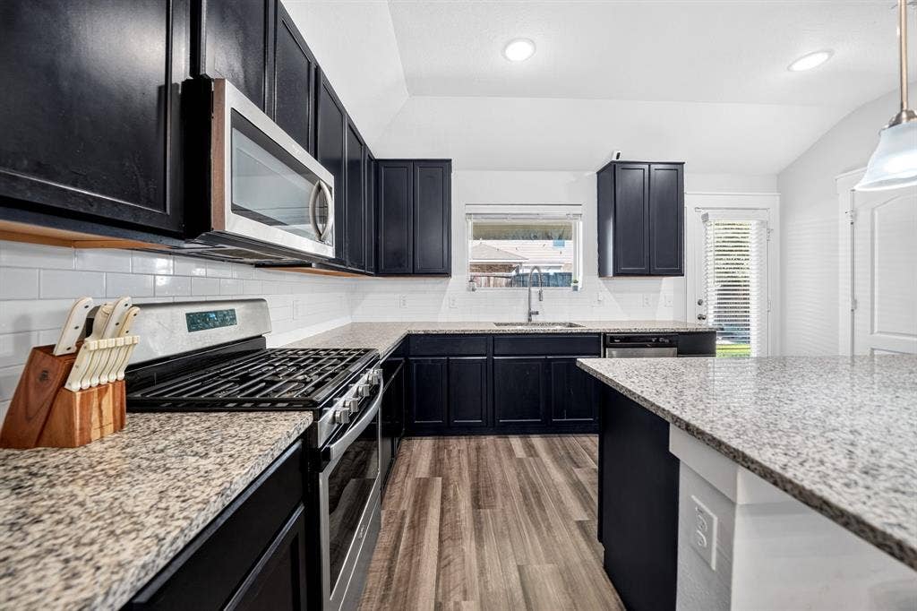 5534 Yarborough Drive, Forney, TX 75126 Modern kitchen featuring sleek black cabinetry with stainless steel gas range, granite-style countertops, subway tile backsplash, recessed ceiling lighting, and wood-finish flooring throughout the open layout