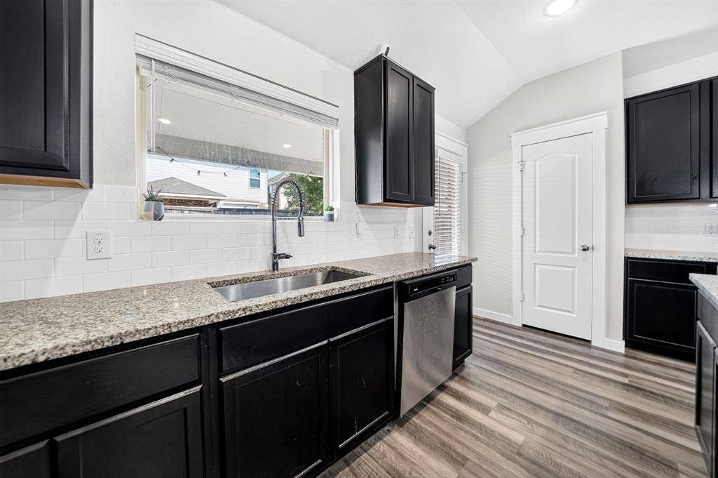 5534 Yarborough Drive, Forney, TX 75126 Modern kitchen featuring sleek black cabinetry with brushed nickel hardware, granite countertops, stainless steel appliances including a professional-style faucet, white subway tile backsplash, and wood-finish plank flooring throughout the space