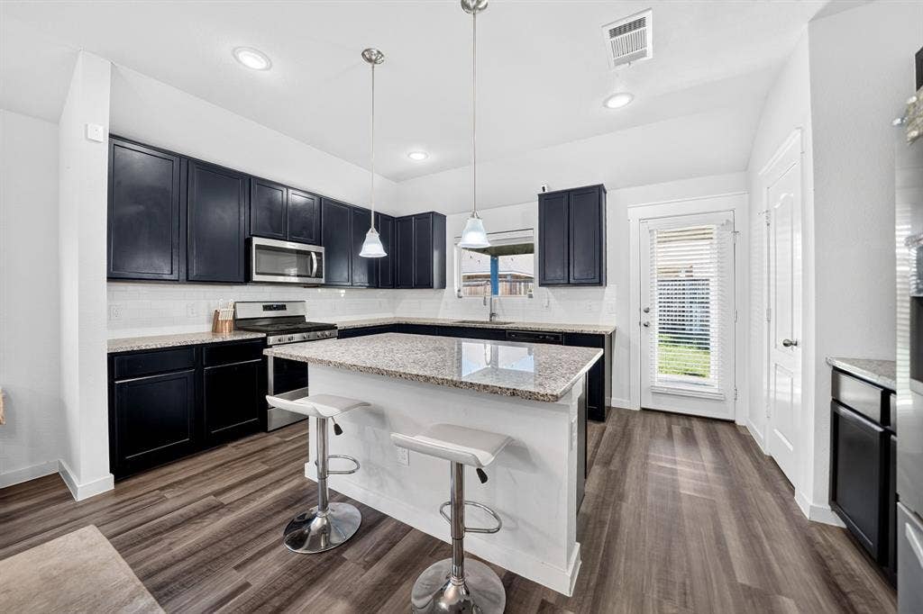 5534 Yarborough Drive, Forney, TX 75126 Modern kitchen featuring dark cabinetry with stainless steel appliances, white quartz countertops, a center island with seating, pendant lighting, and wood-finish flooring throughout the open layout