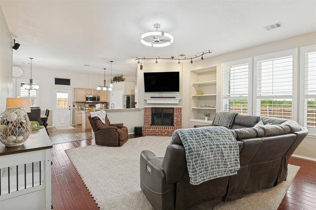 Living area featuring wood-finish flooring, a brick-surround fireplace, built-in shelving, and multiple windows with plantation shutters