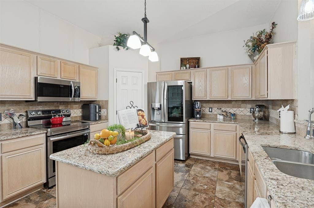 Kitchen featuring a central island, light wood-finish cabinetry, stainless steel appliances, speckled countertops, and a tiled backsplash