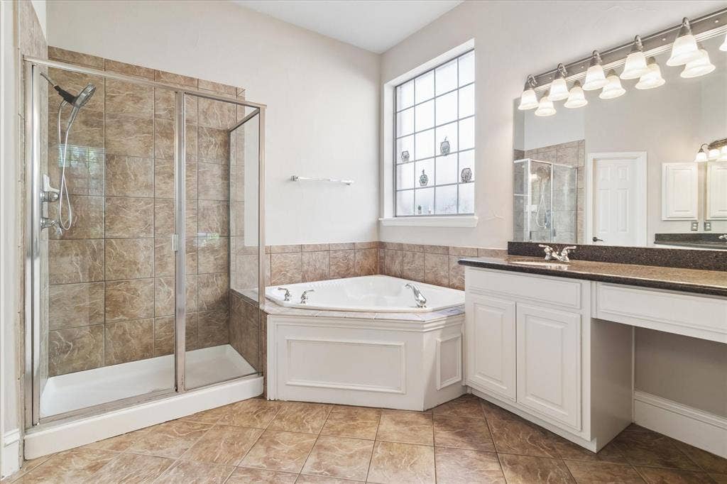 8323 Albritton Drive, Frisco, TX 75036 Spacious bathroom featuring a corner soaking tub, a tiled walk-in shower with glass enclosure, a dual vanity with dark countertops, and a large privacy window