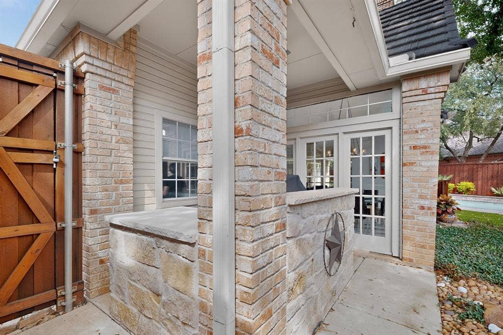 4536 Briar Oaks Circle, Dallas, TX 75287 Covered patio with rusticated brick pillars and stone veneer detailing, complemented by a wooden barn-style gate and mature landscaping along the property perimeter