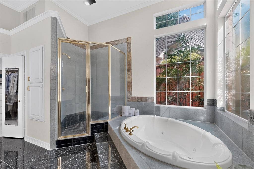 4536 Briar Oaks Circle, Dallas, TX 75287 Soaking tub with brass fixtures, glass-enclosed shower, black polished tile flooring, and transom windows