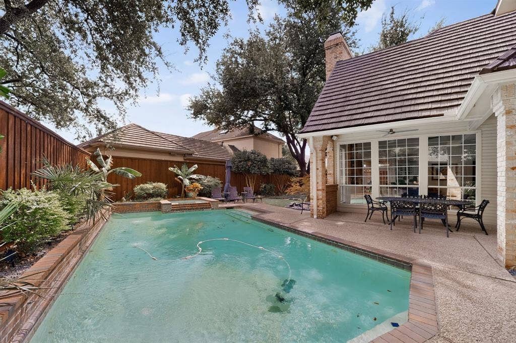 4536 Briar Oaks Circle, Dallas, TX 75287 Resort-style backyard oasis featuring a sparkling turquoise swimming pool with surrounding brick decking, mature shade trees, a covered patio with French doors, and a brick chimney detail on the main residence