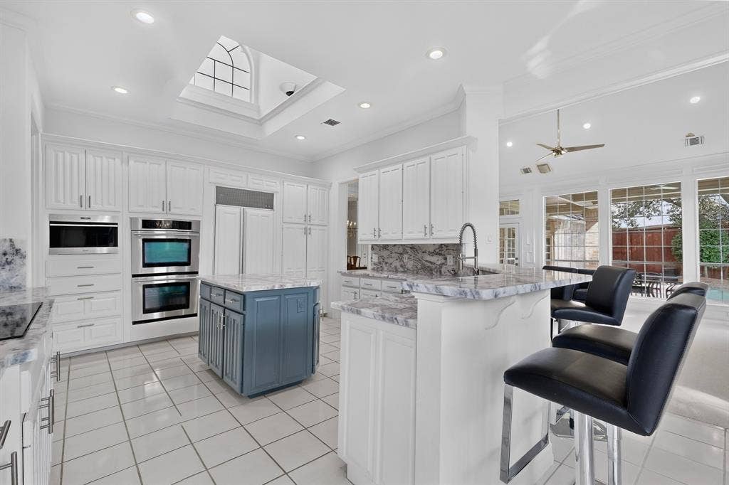4536 Briar Oaks Circle, Dallas, TX 75287 Sophisticated open-concept kitchen and dining area featuring white cabinetry with stainless steel appliances, a dual-island layout with waterfall-edge countertops, tile flooring, recessed and pendant lighting, and direct sightlines to the adjoining living space with large windows providing natural light and outdoor access