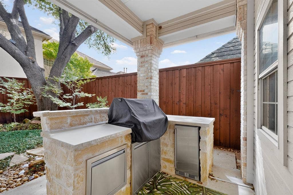 4536 Briar Oaks Circle, Dallas, TX 75287 Covered patio featuring a built-in stone grill with stainless steel appliances and concrete countertop, framed by a mature shade tree and ornamental fencing