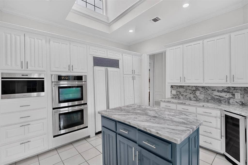 4536 Briar Oaks Circle, Dallas, TX 75287 Chef's kitchen featuring white cabinetry with stainless steel hardware, dual wall-mounted ovens, a blue-gray island with marble-finish countertops, stainless steel appliances, tile flooring, and skylight illumination overhead