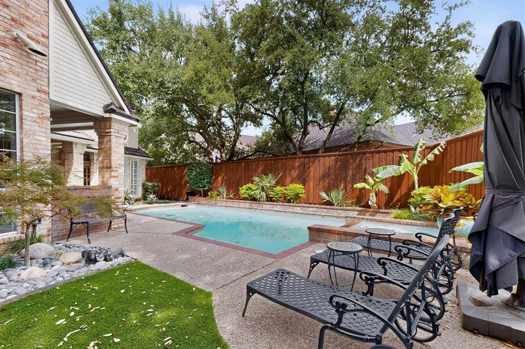 4536 Briar Oaks Circle, Dallas, TX 75287 Expansive backyard oasis featuring a resort-style swimming pool with sun lounging area, mature shade trees, manicured lawn, wood privacy fencing, and brick exterior home with covered patio access