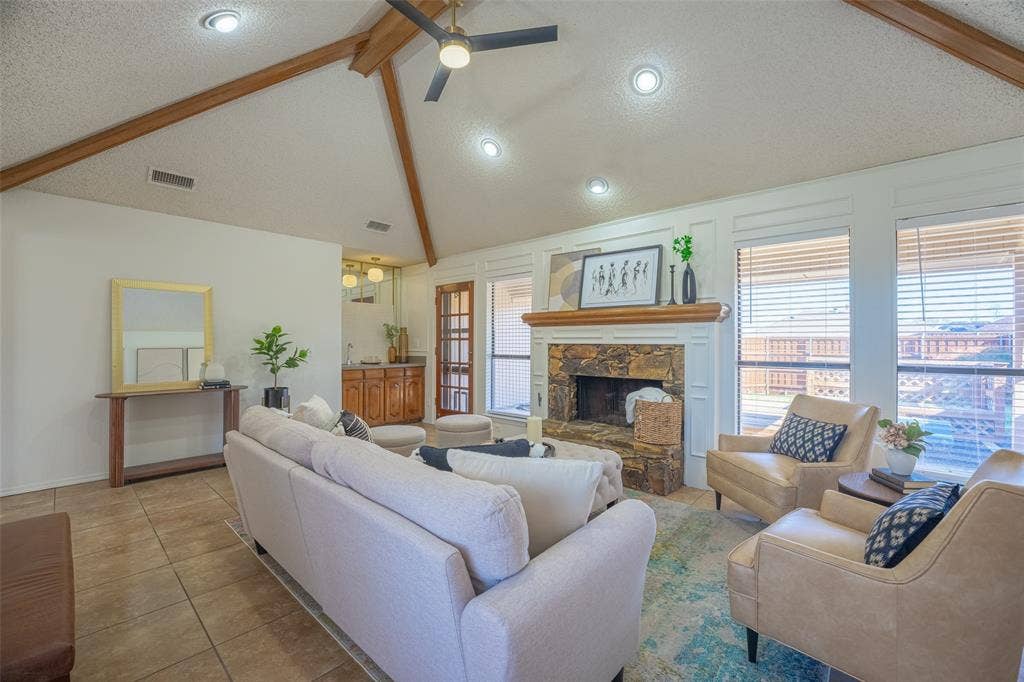 410 W Oak Street, Wylie, TX 75098 Vaulted ceiling with exposed wood beams and recessed lighting, stone-clad fireplace with white mantel, tile flooring, built-in wet bar with wood cabinetry, and multiple windows with blinds