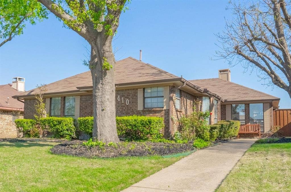 410 W Oak Street, Wylie, TX 75098 Brick exterior with a brown shingle roof, mature front yard tree, established shrubbery, and a concrete pathway