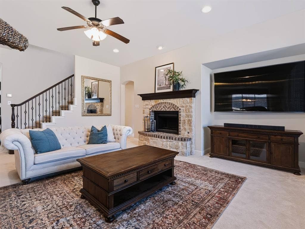 Stone fireplace with dark wood mantel, recessed lighting, ceiling fan, and staircase with wrought iron balusters