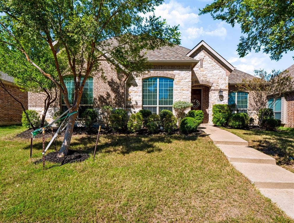 Brick and stone facade, arched entryway, gabled roof, mature landscaping, and concrete pathway