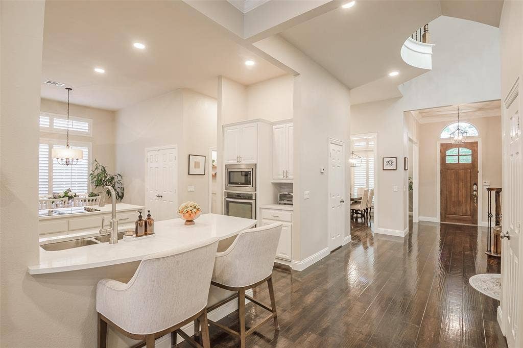 5925 Westmont Drive, Plano, TX 75093 Hardwood-style flooring, a spacious kitchen island with seating, white shaker cabinetry, stainless steel built-in appliances, and a wood-panel entry door with an arched transom