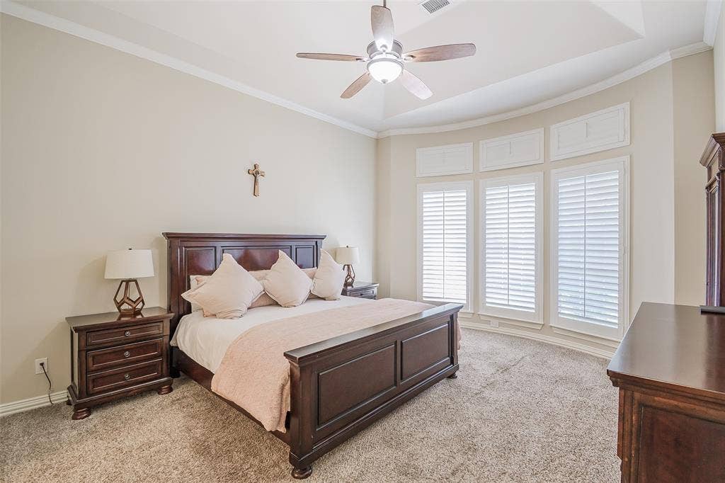 5925 Westmont Drive, Plano, TX 75093 Spacious primary bedroom featuring dark wood architectural detailing, dual nightstands with task lighting, ceiling fan with integrated light fixture, and expansive glass doors with plantation shutters opening to natural light