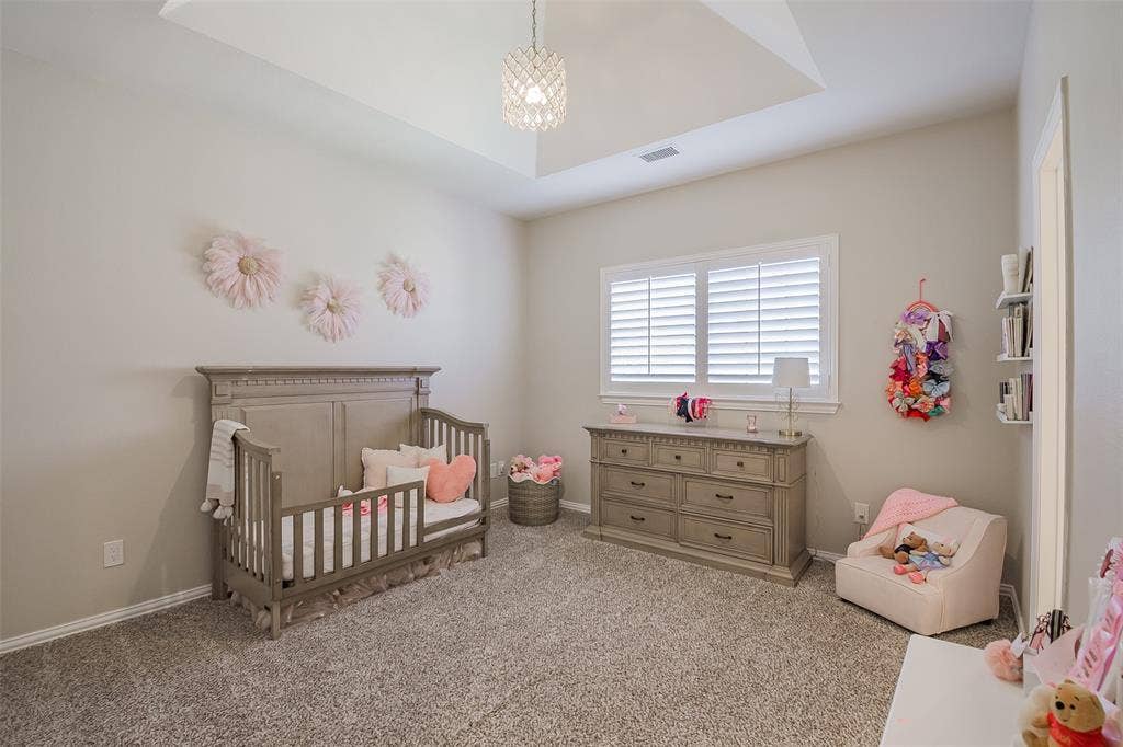 5925 Westmont Drive, Plano, TX 75093 Bright nursery featuring a coffered ceiling with recessed lighting, plantation shutters framing large windows, neutral carpeting, coordinating wood-finish dresser and crib, and soft ambient décor throughout the space