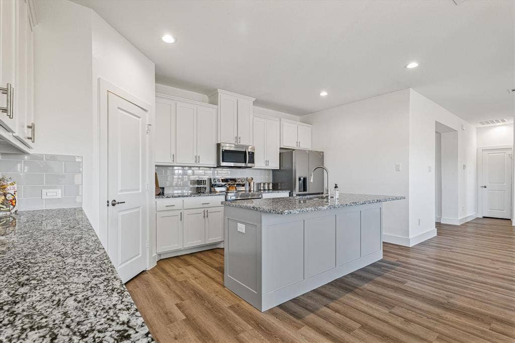Kitchen featuring white cabinetry, light gray subway tile backsplash, stainless steel appliances, a center island with a sink, and wood-finish flooring