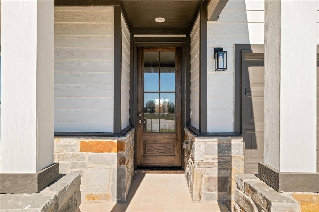 Crafted wood-finish entry door with clear glass panels, stone veneer facade, horizontal siding, and a black sconce light fixture
