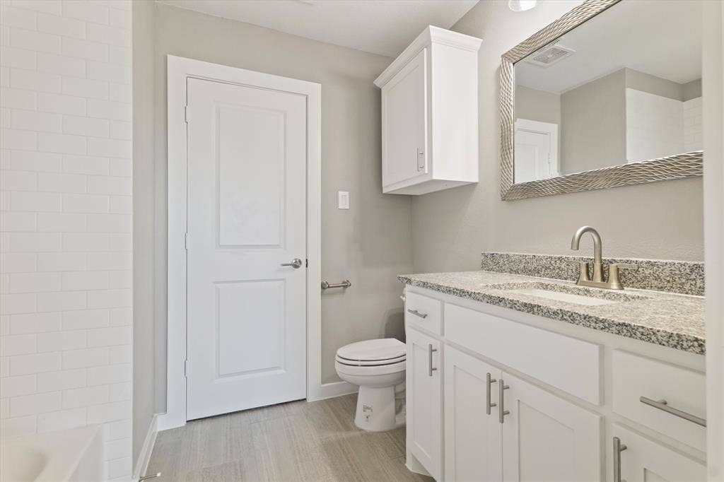 White subway tile surround, wood-finish flooring, white vanity with stone countertop, stainless steel sink with single-handle faucet, and mirrored wall cabinet