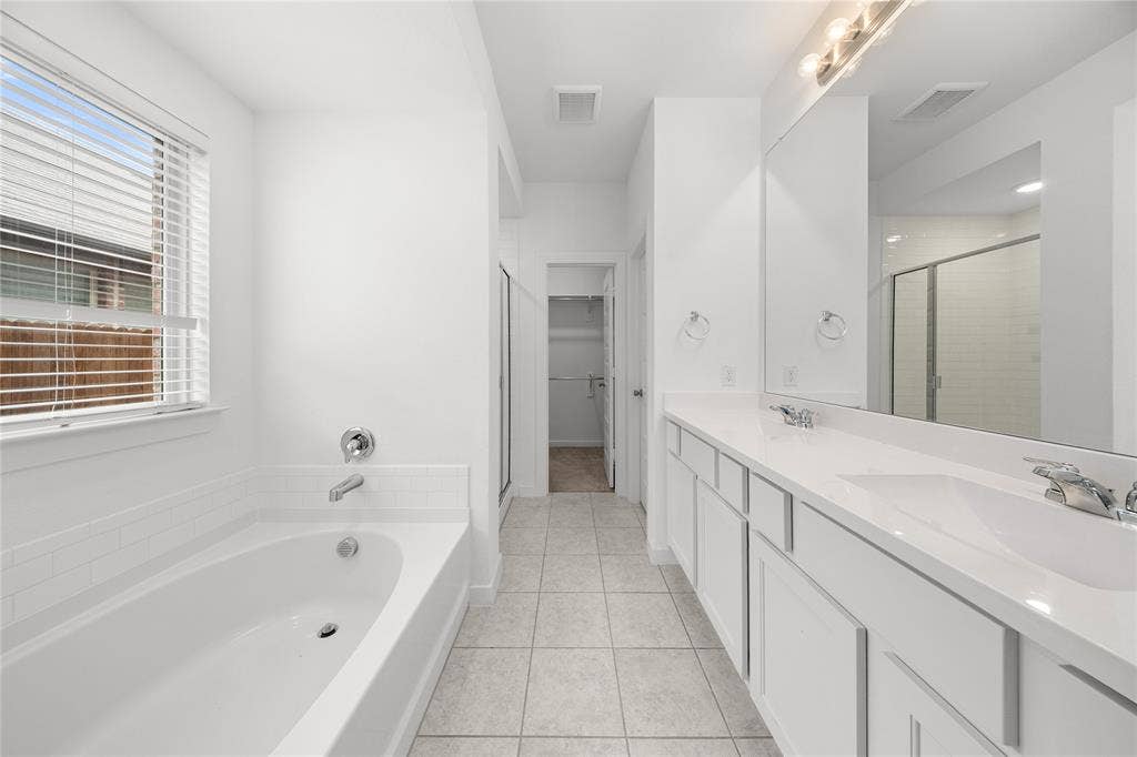 3955 Chesapeake Lane, Heartland, TX 75126 White tile flooring, a built-in bathtub with subway tile surround, a window with horizontal blinds, a vanity with dual integrated sinks, and a framed mirror with overhead lighting