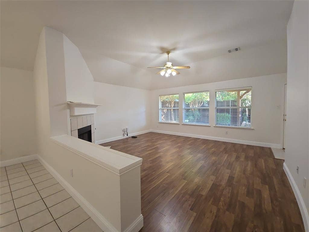 2424 Brycewood Lane, Plano, TX 75025 Open-concept living area featuring a vaulted ceiling with a ceiling fan, wood-finish flooring, a tile-surround fireplace, and three large windows