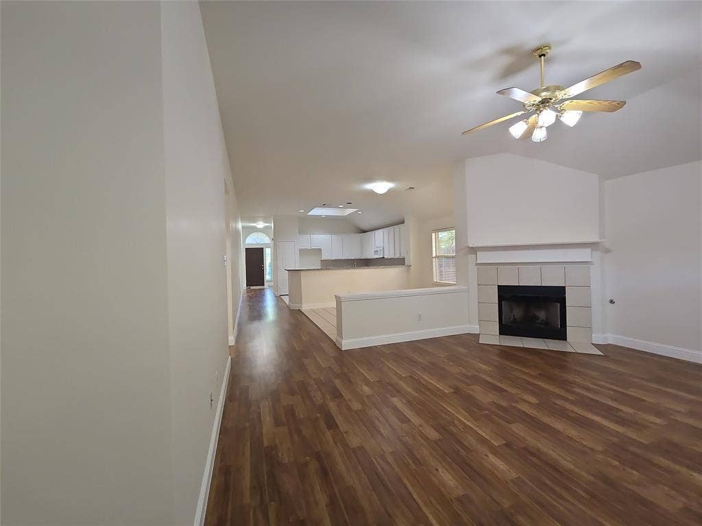 2424 Brycewood Lane, Plano, TX 75025 Vaulted ceiling with ceiling fan, fireplace with tiled surround, wood-finish flooring, and a kitchen with white cabinetry
