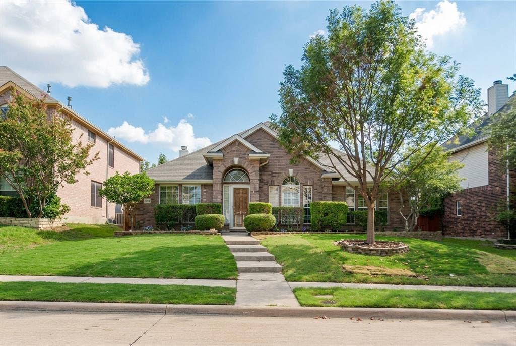 2424 Brycewood Lane, Plano, TX 75025 Brick exterior with an arched entry, manicured landscaping, mature trees, and a concrete walkway