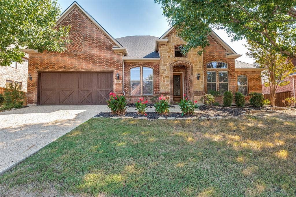 1504 Daisy Lane, Flower Mound, TX 75028 Brick and stone exterior featuring an arched entryway, decorative garage door, and mature landscaping
