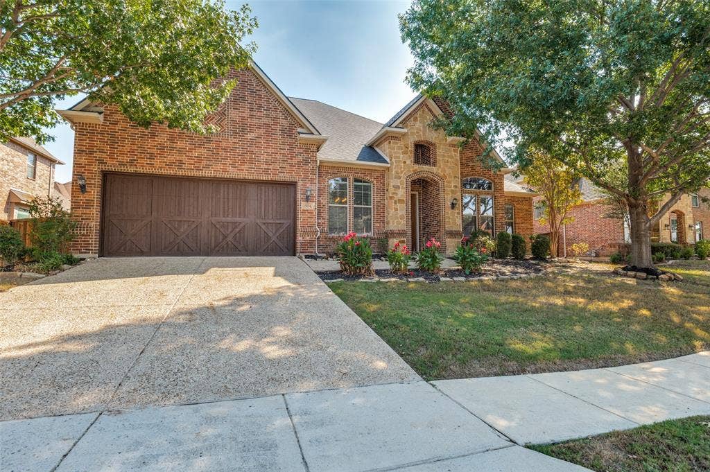 1504 Daisy Lane, Flower Mound, TX 75028 Brick exterior with stone accents, arched entryway, decorative wood garage door, concrete driveway, and established landscaping
