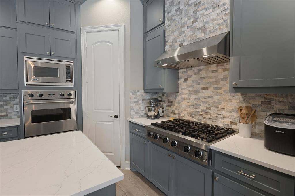 1504 Daisy Lane, Flower Mound, TX 75028 Gray cabinetry, built-in stainless steel oven and microwave, professional-grade gas range with stainless steel hood, quartz countertops, and a stone tile backsplash