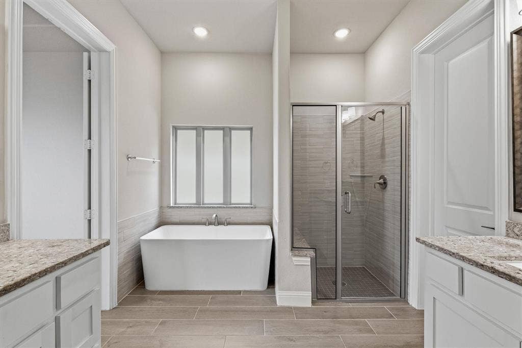 Freestanding bathtub with a tiled surround, separate glass-enclosed shower with a built-in bench, dual vanities with stone countertops, and wood-finish flooring