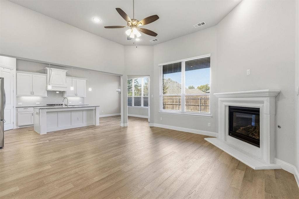 Open-concept living space featuring wood-finish flooring, a white cabinetry kitchen with stainless steel appliances, a fireplace with white surround, ceiling fan with integrated lighting, multiple large windows with views of exterior landscapes, and glass sliding doors providing access to an outdoor deck