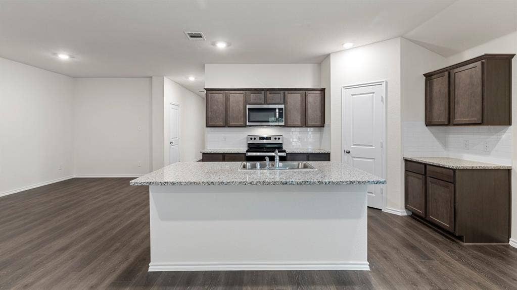 5012 Shumard Drive, Lindale, TX 75771 Kitchen featuring a center island with a built-in sink, stainless steel appliances, dark wood-finish cabinetry, light-colored countertops, and white subway tile backsplash