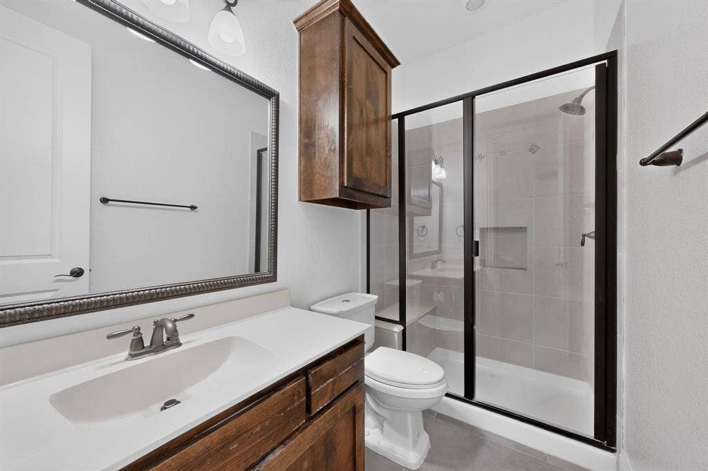 13853 Myatt Avenue, Frisco, TX 75035 Bathroom featuring a spacious walk-in shower with clear glass enclosure and built-in niche, a single vanity with an integrated sink and brushed nickel faucet, a large framed mirror, a wall-mounted wooden storage cabinet, and white walls