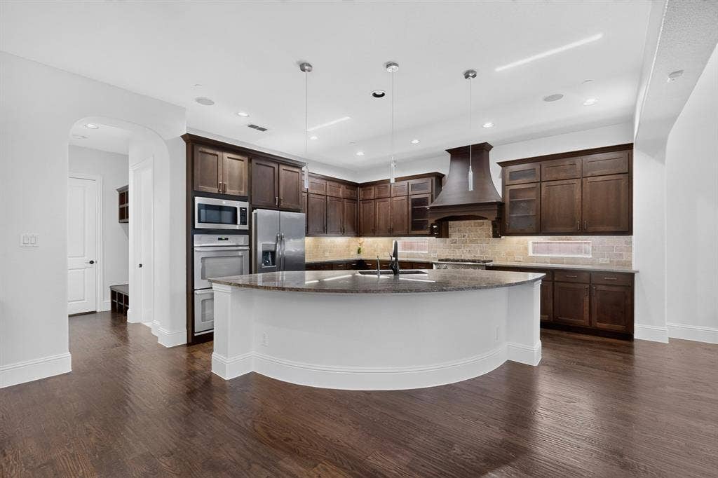 13853 Myatt Avenue, Frisco, TX 75035 Spacious kitchen featuring an expansive curved island with a stone countertop, dark wood-finish cabinetry, stainless steel appliances, and wood-finish flooring