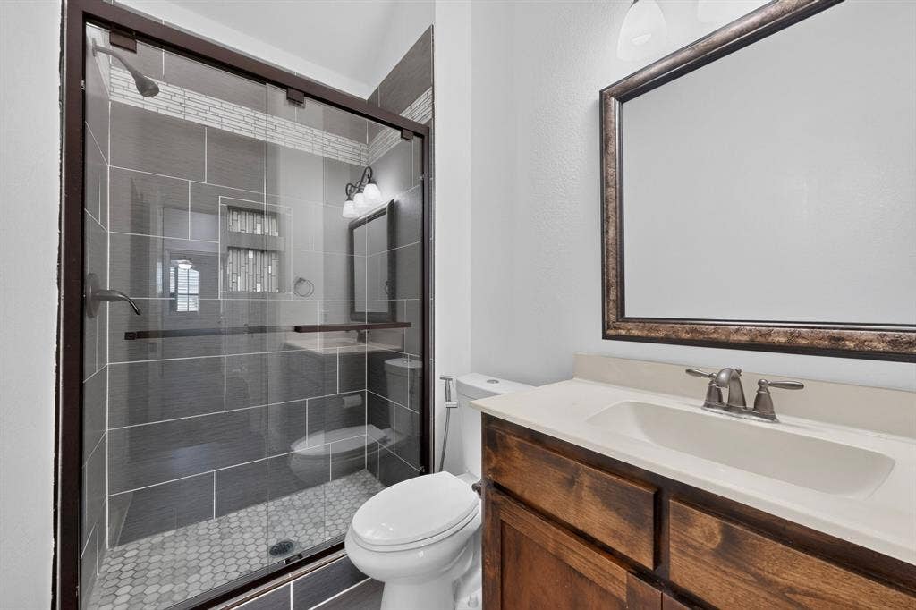 13853 Myatt Avenue, Frisco, TX 75035 Glass-enclosed shower with dark tile surround and mosaic floor, wood-finish vanity with an integrated sink, framed mirror, and brushed nickel fixtures