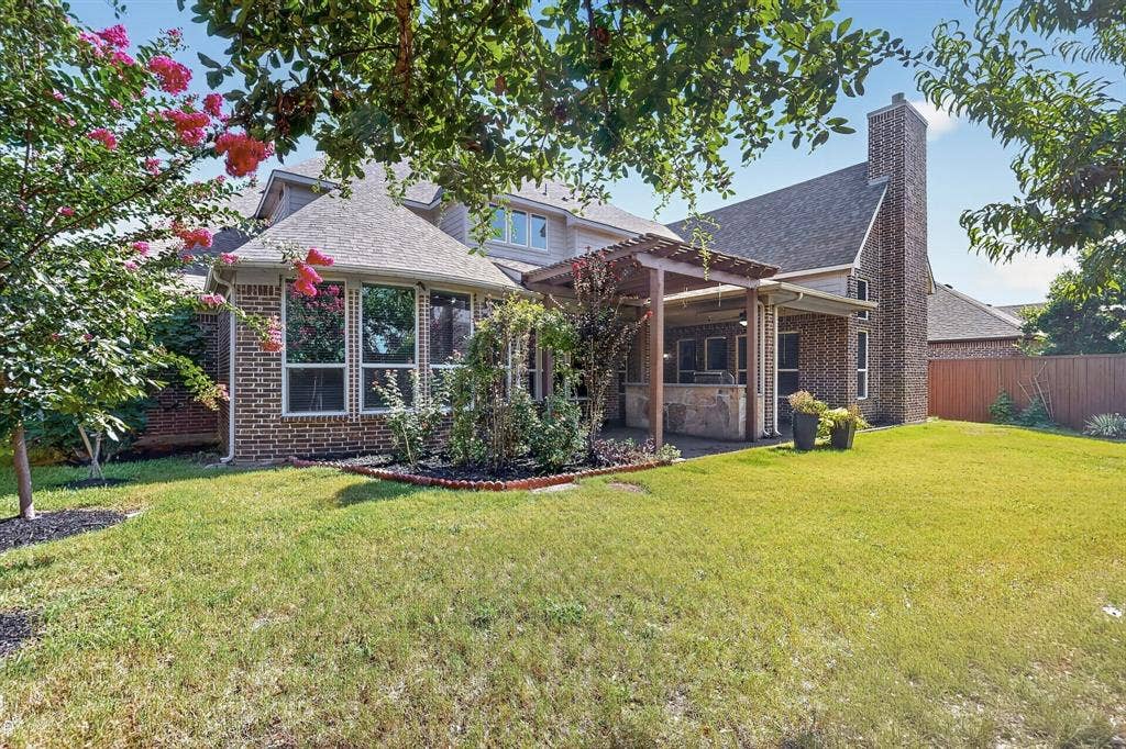 13853 Myatt Avenue, Frisco, TX 75035 Expansive backyard featuring a manicured lawn, a covered patio with a built-in outdoor kitchen, an attached pergola, a brick chimney, and a sunroom with multiple windows