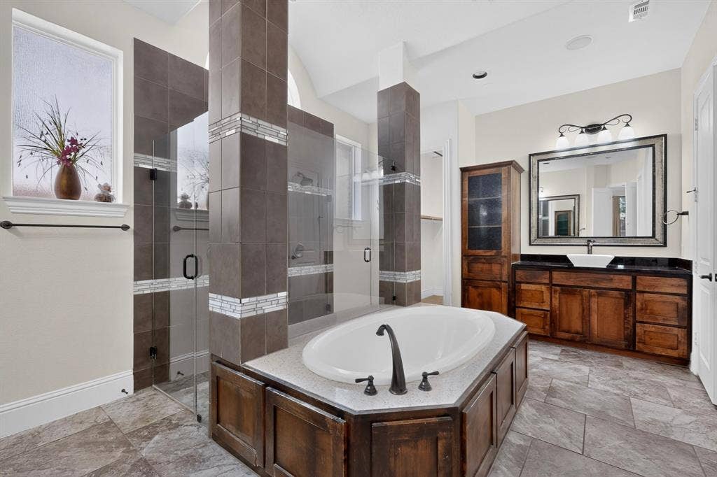 13853 Myatt Avenue, Frisco, TX 75035 Spacious bathroom featuring a built-in soaking tub, a tiled walk-in shower with glass enclosures, a wood-finish vanity with an undermount sink, a large framed mirror, and tile flooring