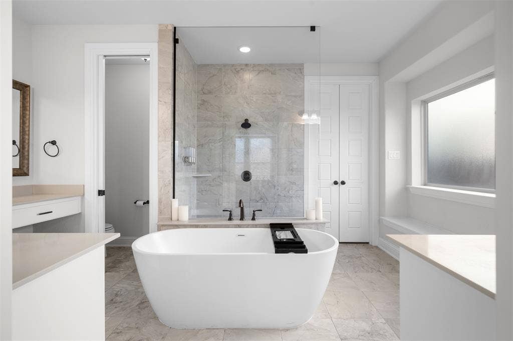Freestanding soaking tub, walk-in shower with glass enclosure, dual vanity with undermount sinks, tile flooring, and a large window with a built-in bench