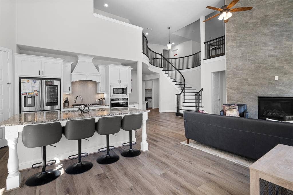 Open concept living space featuring wood-finish flooring, a kitchen island with stone countertop, stainless steel appliances, a curved staircase with black metal balusters, and a fireplace with a full-height tile surround