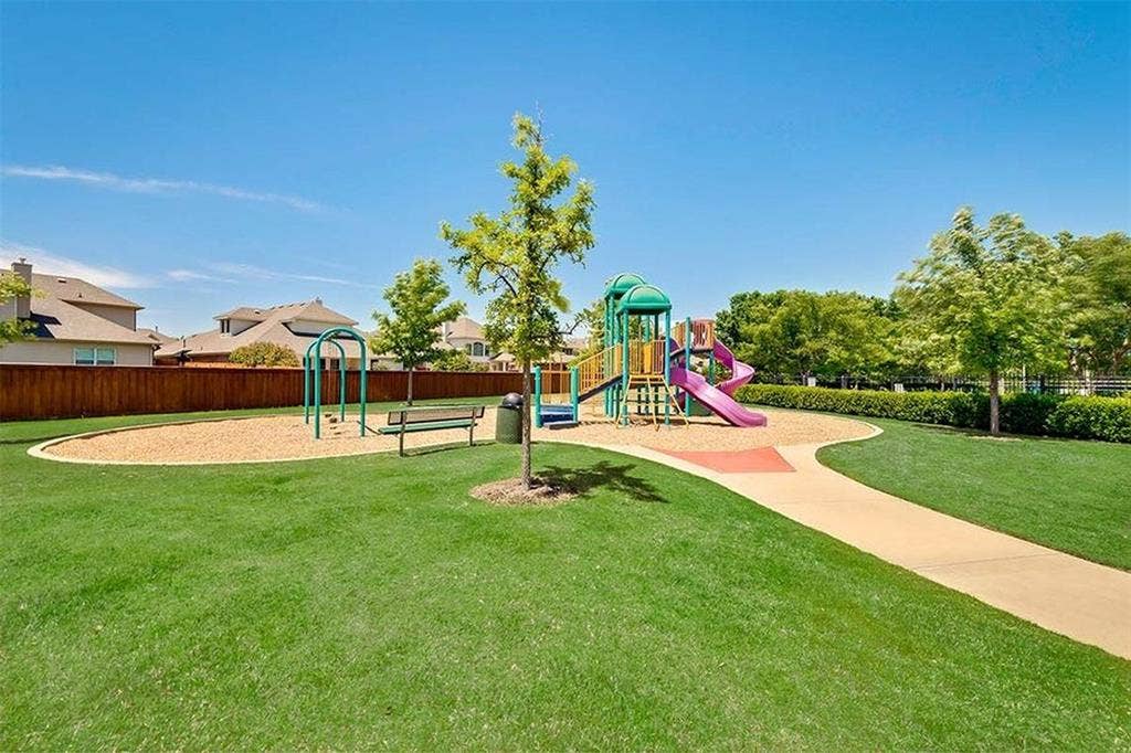 Expansive green space featuring a playground with slides and swings, a paved pathway, and a perimeter of mature trees