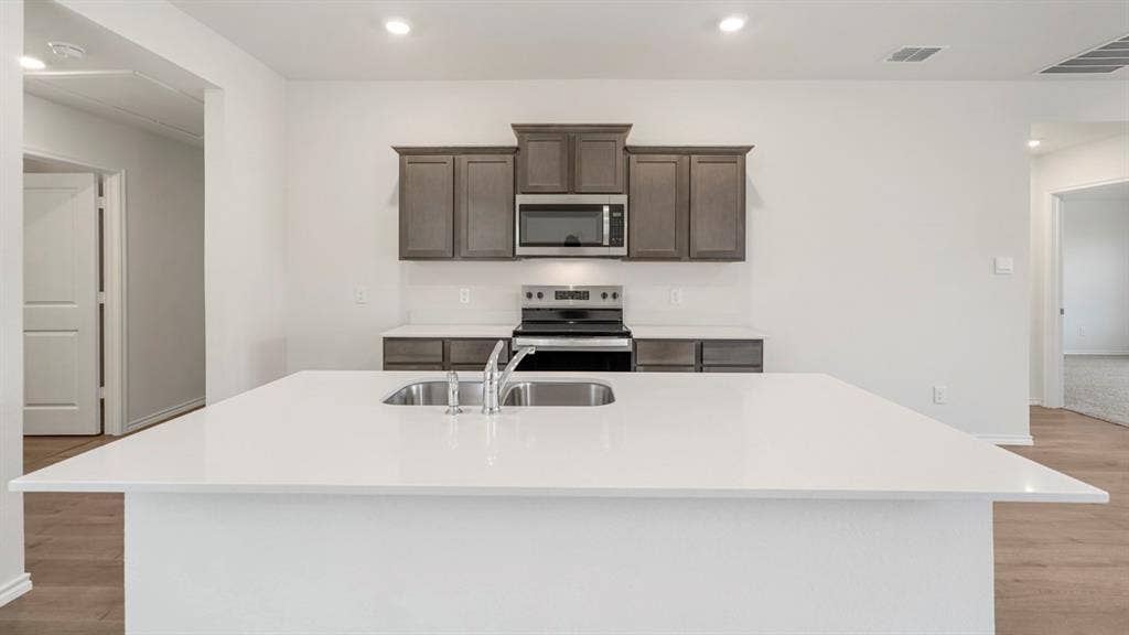 Kitchen featuring an expansive island with a double basin stainless steel sink, light-colored countertops, wood-finish flooring, recessed lighting, and dark wood cabinetry with stainless steel appliances