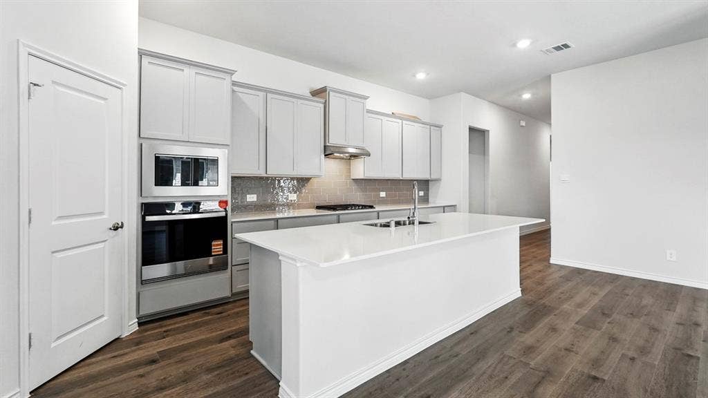 2503 Wesley Avenue, Royse City, TX 75189 Modern kitchen featuring a large central island with an undermount sink, light gray cabinetry, stainless steel appliances, a tiled backsplash, and wood-finish flooring